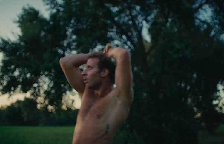 Shirtless man performs a dynamic steel mace workout in a natural outdoor setting at dusk