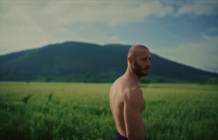 A muscular man turns to the camera before his friend joins him to look at the mountain view from a green field