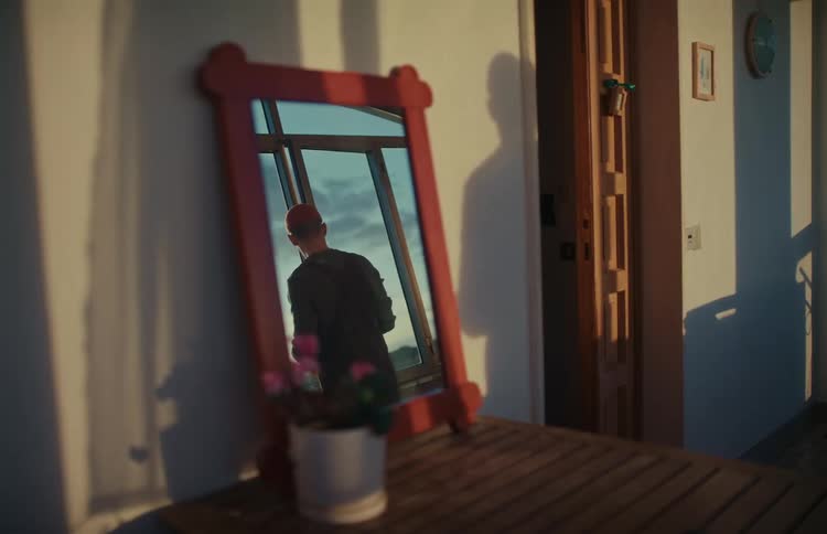 A red-framed mirror on a balcony reflects a man cleaning a window as a shadow is cast on the wall at sunset
