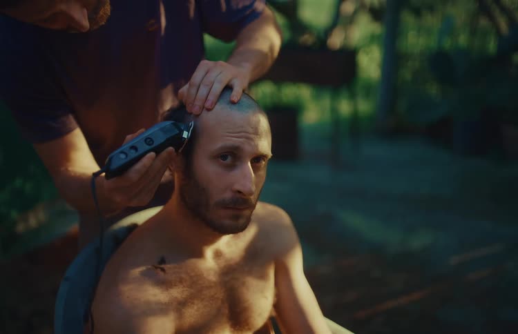 A serious man looks into the camera while another shaves his head with clippers in a rustic outdoor setting at dusk