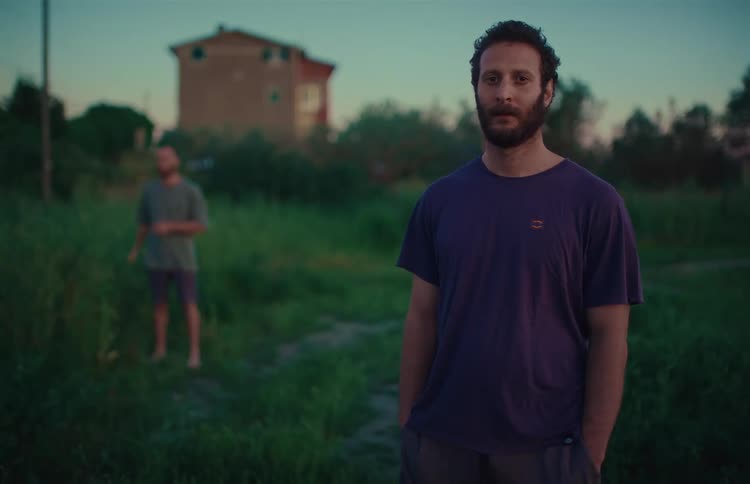 A cinematic portrait of a serious young man with a beard standing in a rural field at dusk