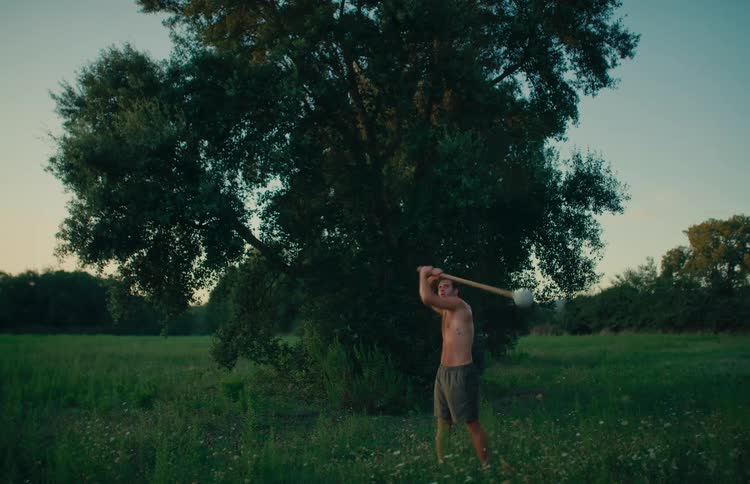 A strong shirtless man performs a steel mace 360 exercise in a green field at sunset