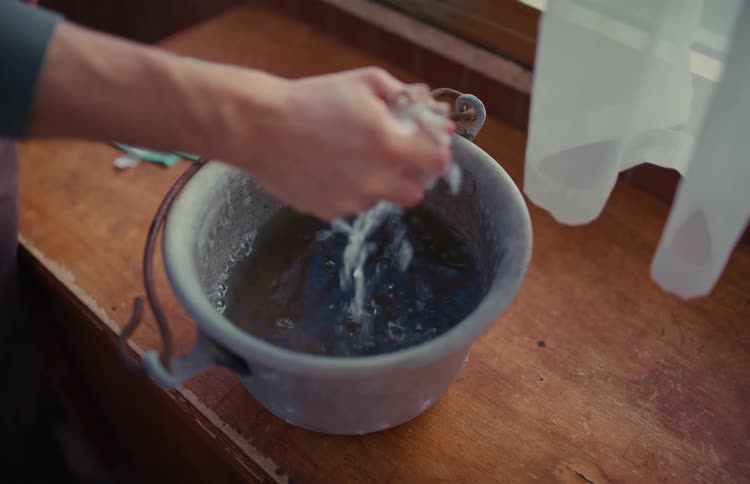 A person's hands slowly wring out a soapy cloth over an old metal bucket of water