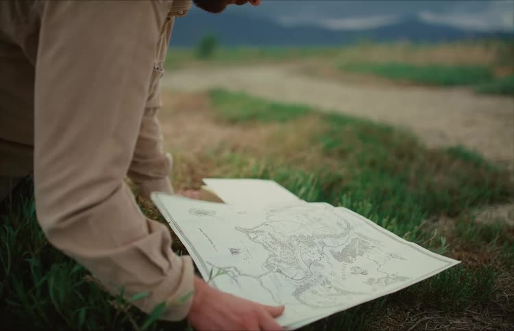 An explorer kneels in a grassy field and carefully studies a vintage fantasy map to plan his journey