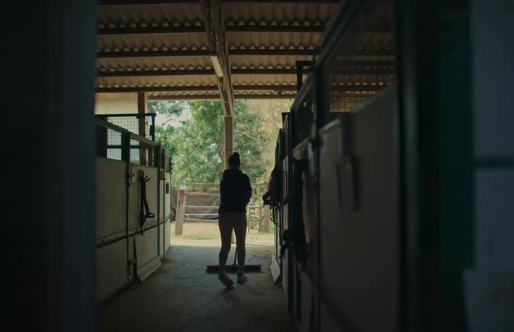 A woman sweeps the floor of a dimly lit aisle in a horse stable during the day