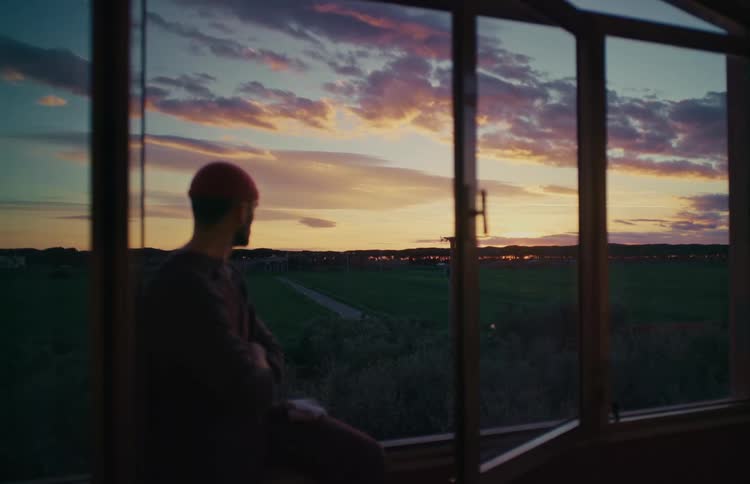 A contemplative man sits by a window and looks out at the stunning, colorful sunset over a rural landscape