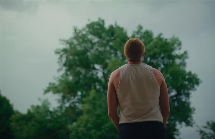 Low-angle shot from behind a man in a beanie and tank top as he stands and looks at a large tree under a cloudy sky