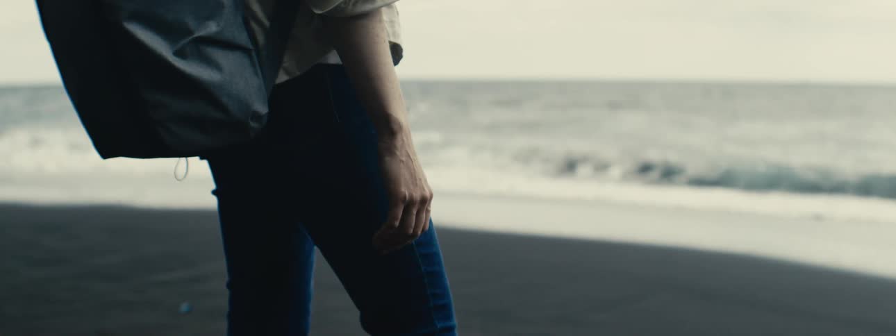 A person with a backpack stands on a black sand beach watching the slow-motion ocean waves