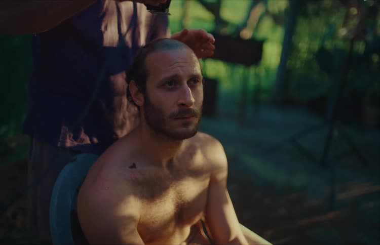 A shirtless man has his head shaved by another man using an electric clipper in a rustic outdoor setting at dusk