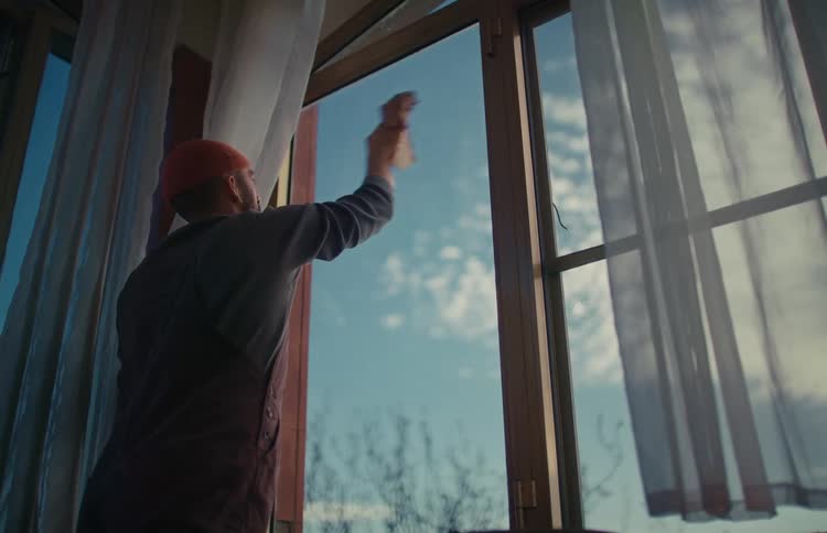 A man in overalls and a red beanie diligently cleans a large window from inside a room on a sunny day