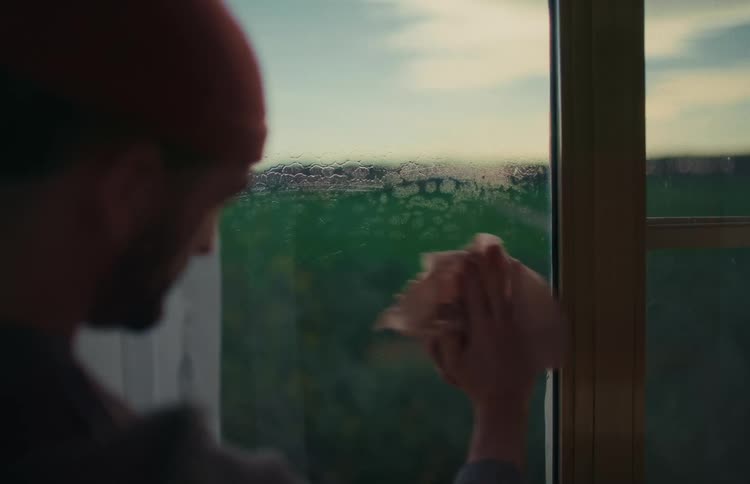A man in a red beanie sprays and wipes a window clean with a cloth, looking out at a green landscape