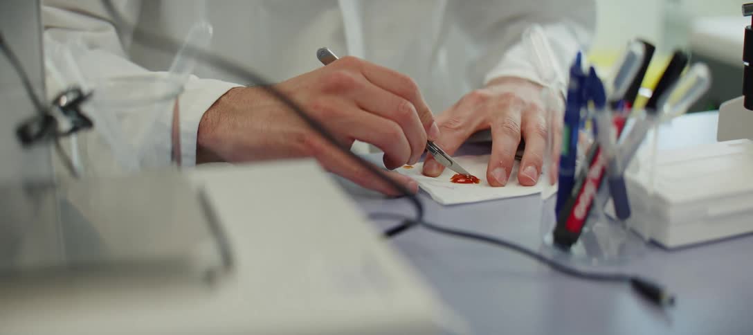 A scientist carefully dissects a small reddish-brown sample with a scalpel on a piece of paper in a sterile laboratory setting