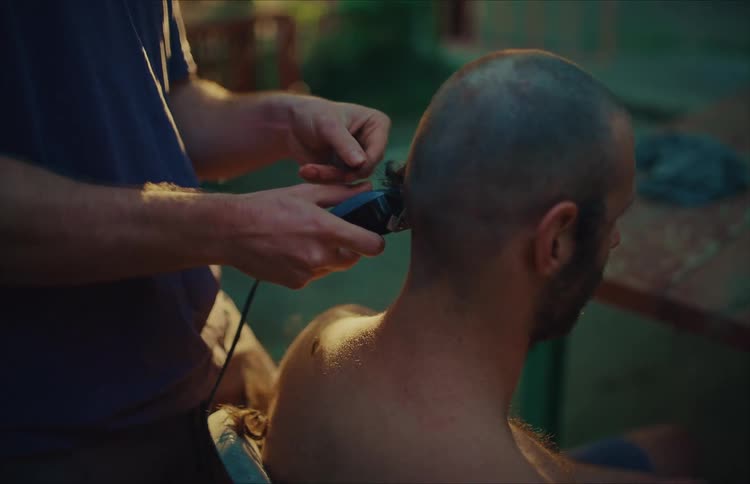 A man shaves another man's head with electric clippers in a backyard during sunset, shot in slow motion