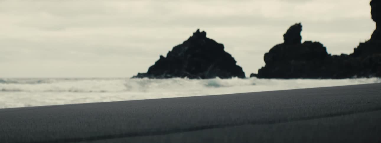 Gentle waves wash ashore on a desolate black sand beach with dramatic sea stacks in the distance