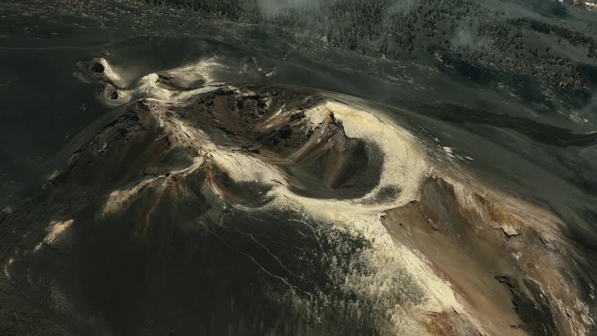Expansive aerial shot slowly pulls away from a dormant volcanic crater, revealing the stark, ash-covered landscape and forest
