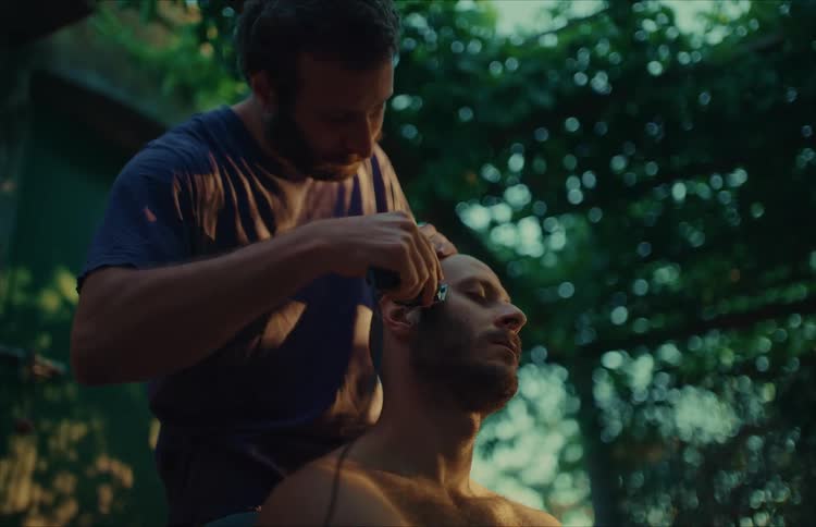 A man shaves his friend's head with an electric clipper in a quiet, sun-dappled backyard