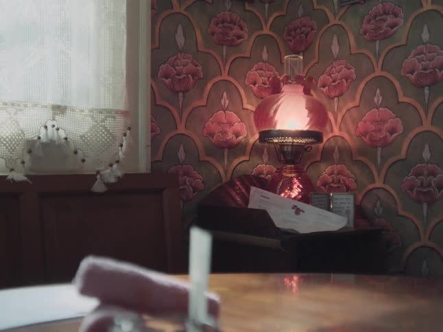 A vintage pink glass oil lamp glows warmly in a cozy room with floral wallpaper next to a sunlit window with a lace curtain