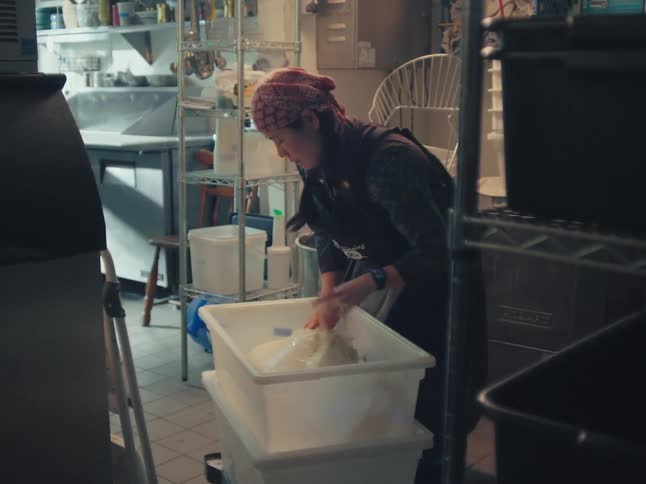 A dedicated baker carefully kneads a large batch of fresh dough by hand in a dimly lit professional kitchen