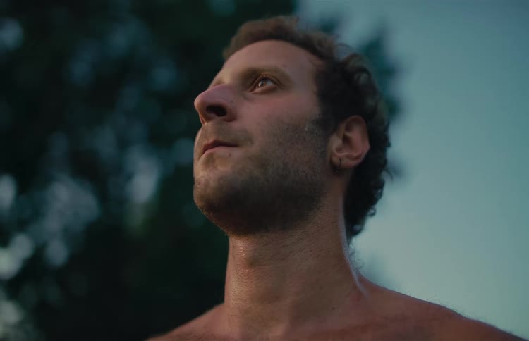 A shirtless man takes a deep breath and opens his eyes, enjoying a moment of peace outdoors at dusk