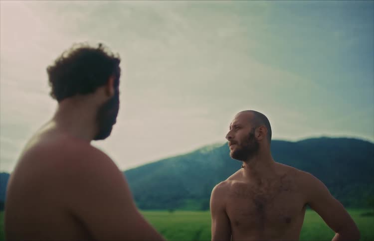 A shirtless bearded man smokes a cigarette and talks to his friend in a scenic mountain landscape
