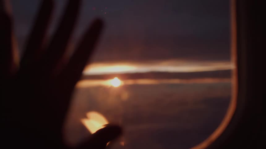 A passenger's hand gently touches the airplane window, reaching towards the golden sunset over a sea of clouds