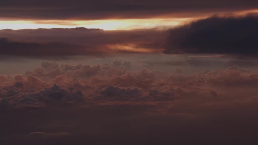 A dramatic and moody aerial view shows a sea of dark, puffy clouds illuminated by the golden light of sunset on the horizon