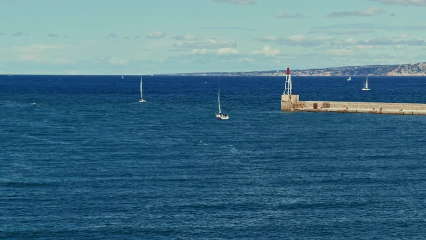 A scenic view of sailboats gliding on the vast blue sea near a stone breakwater with a small lighthouse