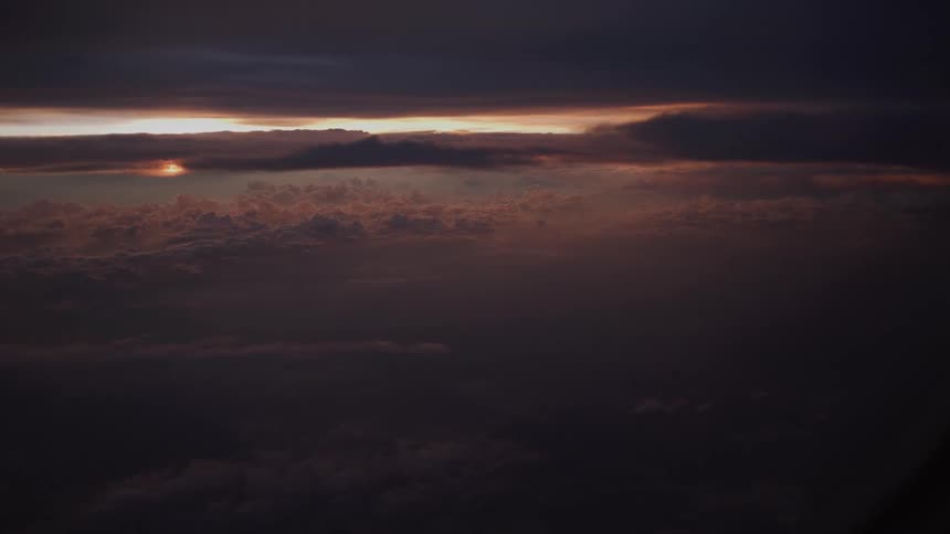 A serene aerial view from an airplane captures the moody sunset peeking through a thick, dramatic layer of clouds