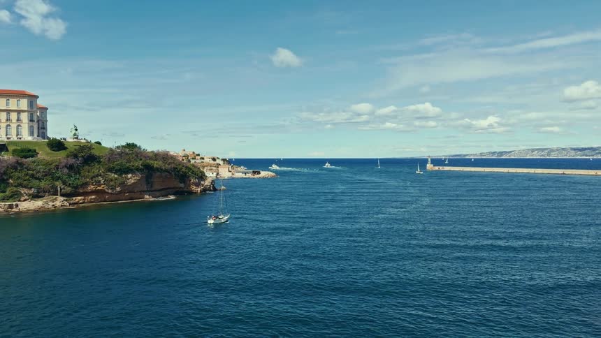 A scenic wide shot of sailboats navigating the deep blue waters at the entrance of a historic European harbor