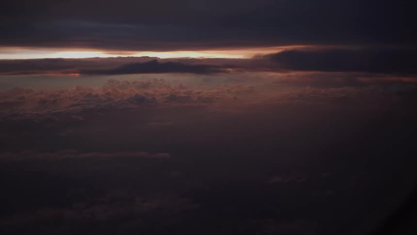 A dramatic aerial view from an airplane window captures a sea of dark clouds illuminated by the last light of a sunset on the horizon