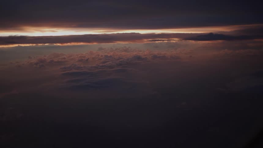A serene and moody aerial view from an airplane window shows a vast cloudscape illuminated by the warm glow of a distant sunset