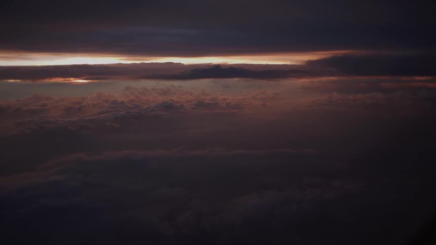 A dramatic aerial shot captures a sea of dark clouds with a thin line of vibrant sunset glowing on the distant horizon