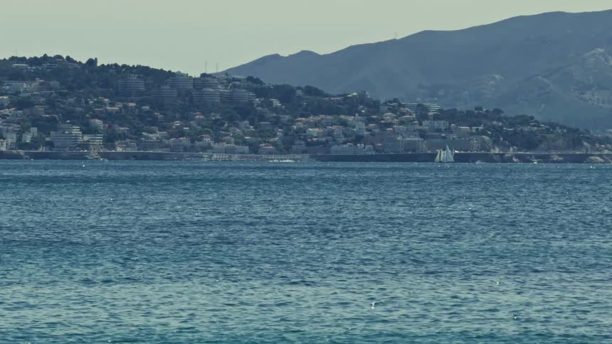 A scenic, static view of the calm blue sea with a distant coastal city nestled on a verdant hillside