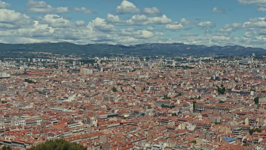 A stunning panoramic view of the sprawling city of Marseille, France, with its iconic red-tiled roofs stretching towards distant mountains under a blue sky