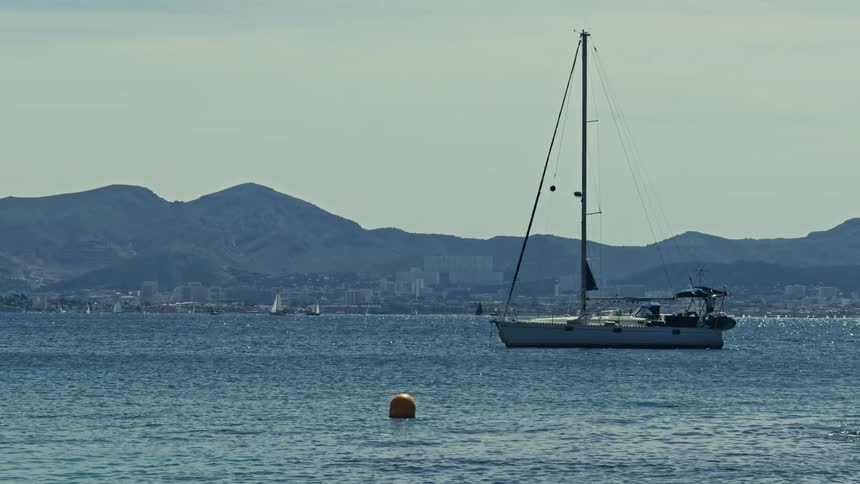 A lone sailboat is anchored in a calm bay with a distant city and mountains on the horizon under a hazy sky