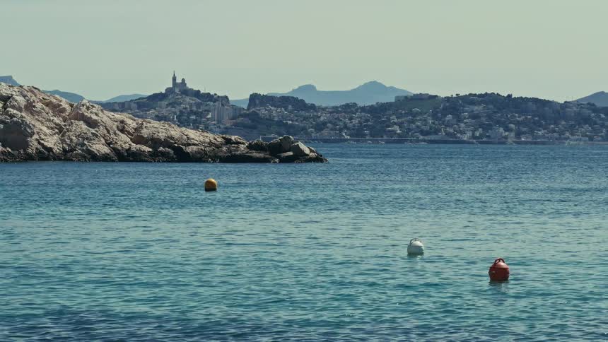 Colorful buoys float on the calm Mediterranean Sea with the iconic Notre-Dame de la Garde and the city of Marseille in the distance