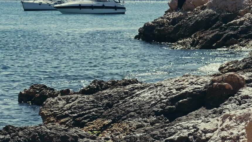 A serene view of the sparkling sea meeting a rugged, rocky shore, with white boats peacefully anchored in the bay