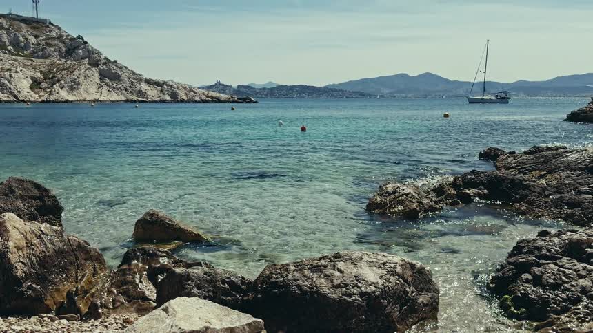A slow tilt-up shot reveals a stunning view of a tranquil turquoise bay from a rocky shoreline with a sailboat and distant mountains