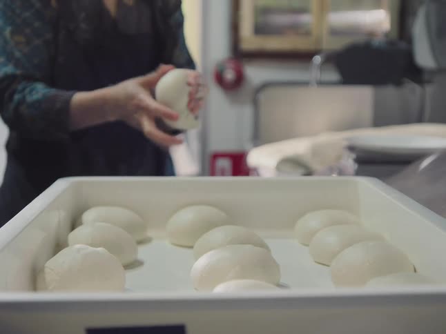 A chef kneads and forms individual pizza dough balls before placing them into a white proofing tray in a commercial kitchen