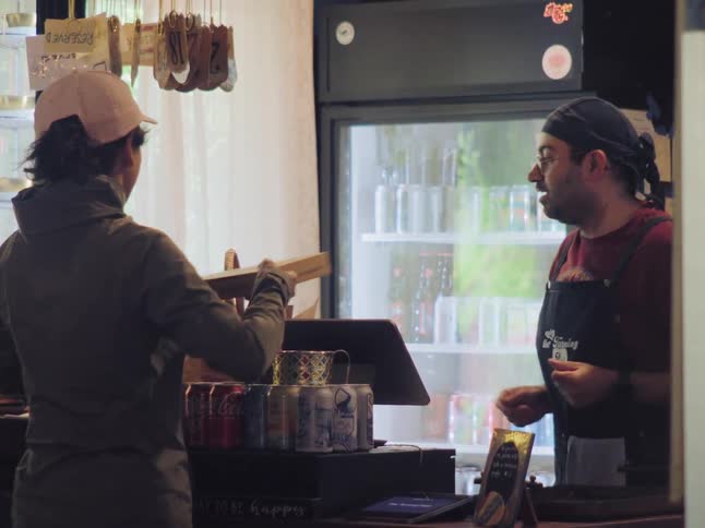 A woman happily receives her takeout pizza and baguette from a friendly employee at a small eatery