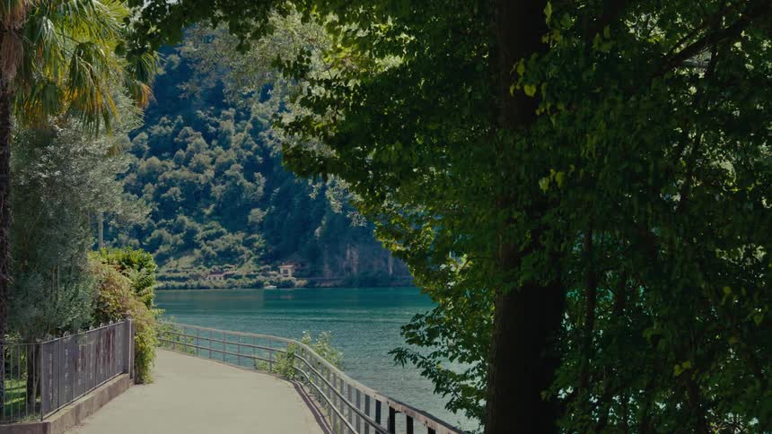 A tranquil paved pathway winds along a serene lake, shaded by lush trees and overlooking a forested mountain