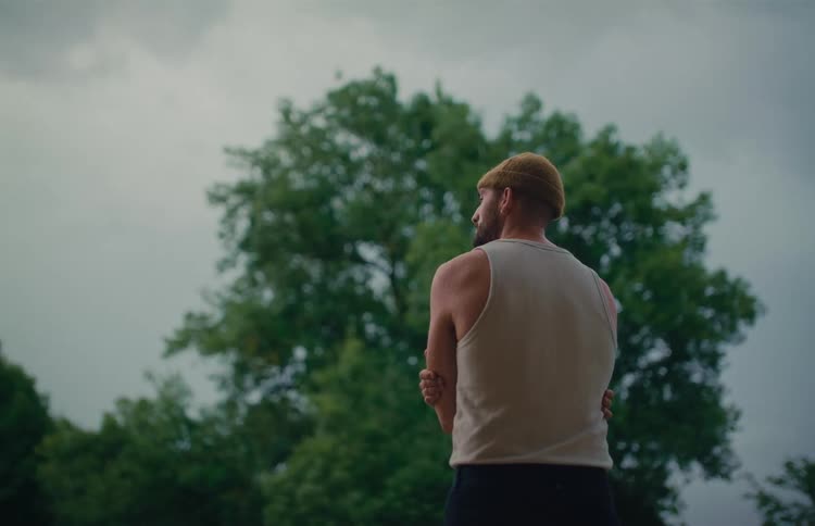 A pensive man in a tank top and beanie stands still with his back to the camera, looking out at a large tree under an overcast sky