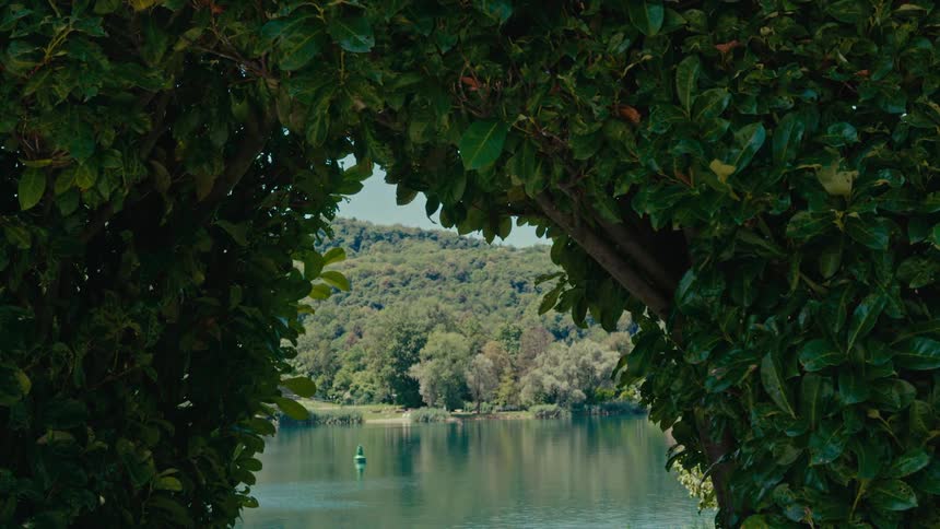A serene lake and a lush, forested hill are viewed through a natural archway of green leaves on a sunny day