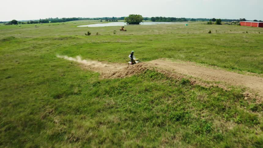 An aerial slow-motion shot captures a motocross rider making a jump on a dirt track in a wide-open grassy field