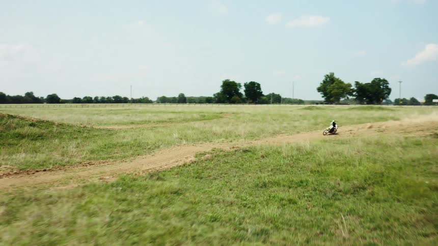 A motocross rider speeds along a dirt track in a large grassy field, kicking up a cloud of dust