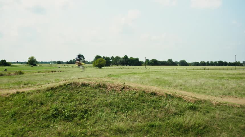 Slow motion aerial shot of a motocross rider launching off a dirt jump in a vast green field