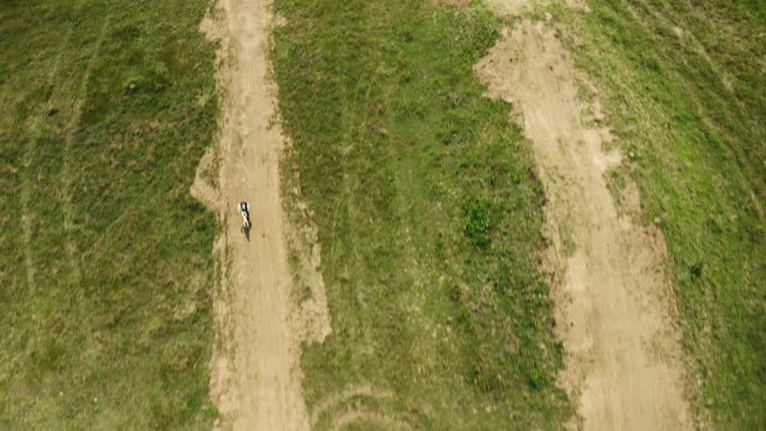 An aerial top-down shot follows a lone cyclist as they navigate a winding dirt trail through a lush green field