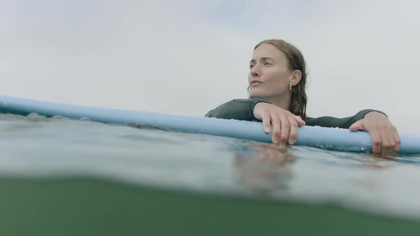 A serene woman in a wetsuit holds onto her surfboard, patiently waiting in the ocean as the camera dips underwater