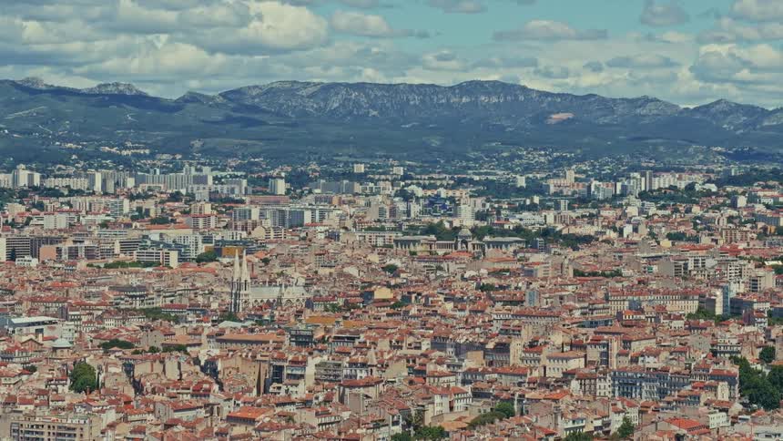A high-angle, wide shot captures the dense, sprawling cityscape of a European city with mountains in the background under a blue sky with white clouds