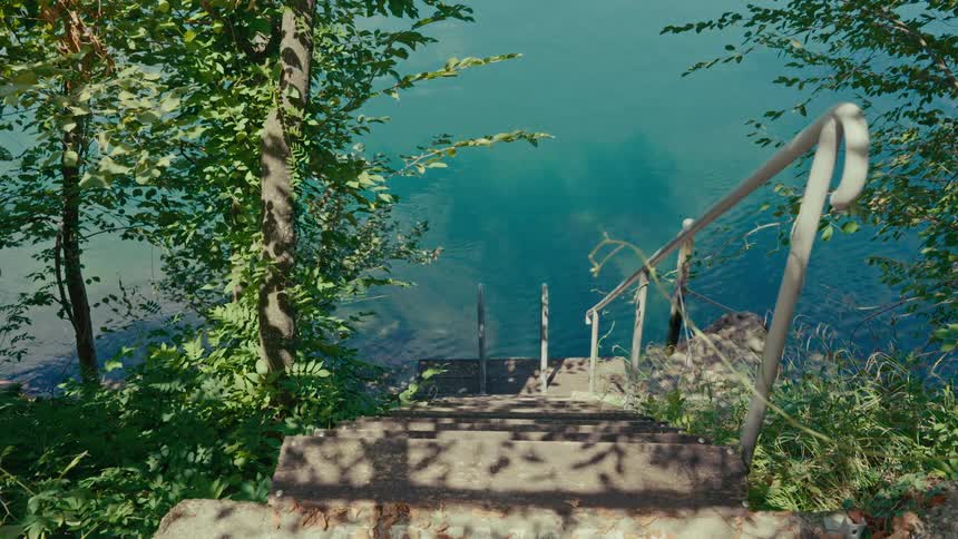 Sun-dappled concrete stairs descend through a green forest to a tranquil, turquoise lake with a handrail for swimming